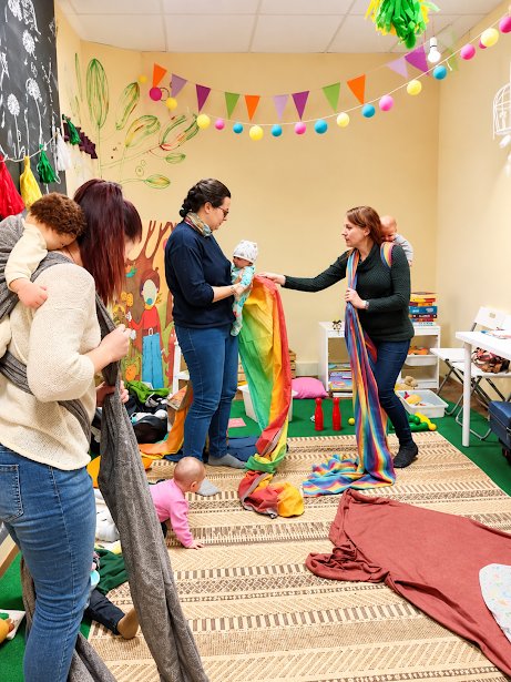 Babahordozós workshop: anyukák hordozókendőket próbálnak ki a babáikkal egy színes, játékos teremben