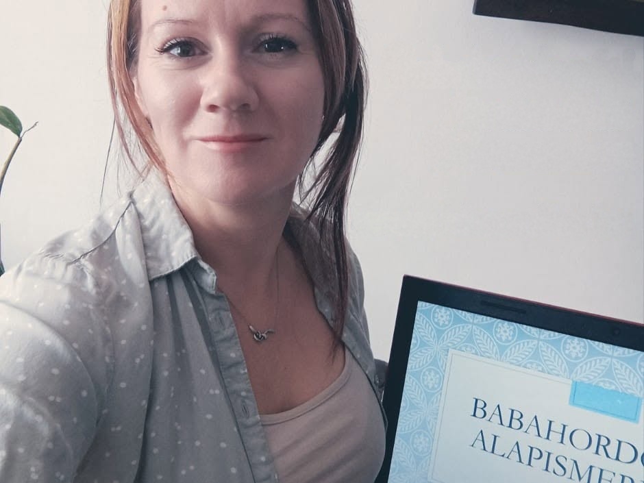Online babahordozási tanácsadás: Kiss Anikó mosolyogva ül a laptopja előtt, a képernyőn a Babahordozás alapismeretek című előadás látszik